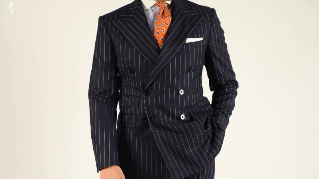 Un costume à double boutonnage bleu marine à fines rayures associé à une pochette de costume blanche et une cravate à motifs orange.