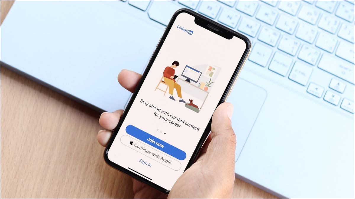 La main d'une personne tenant un smartphone au-dessus d'un clavier d'ordinateur portable, avec l'application LinkedIn à l'écran.