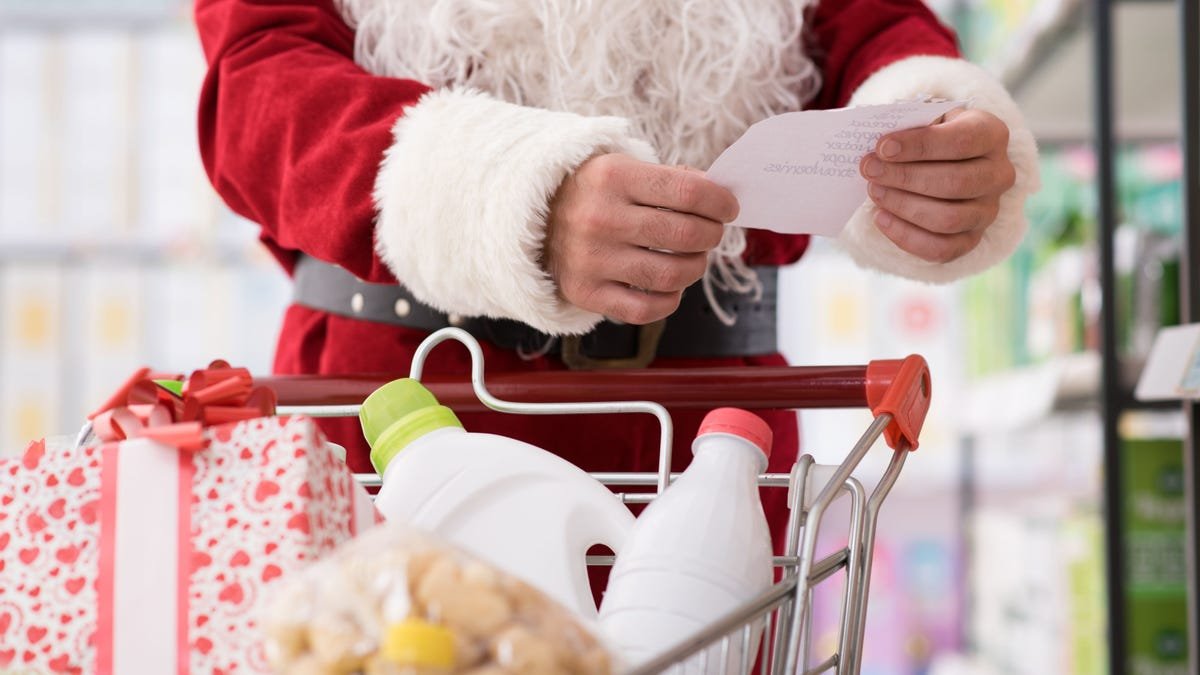 Un Père Noël regarde une liste d'épicerie devant un chariot rempli de provisions.