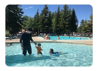 Un père tient la main de son fils dans une piscine et sa fille nage à côté d'eux.