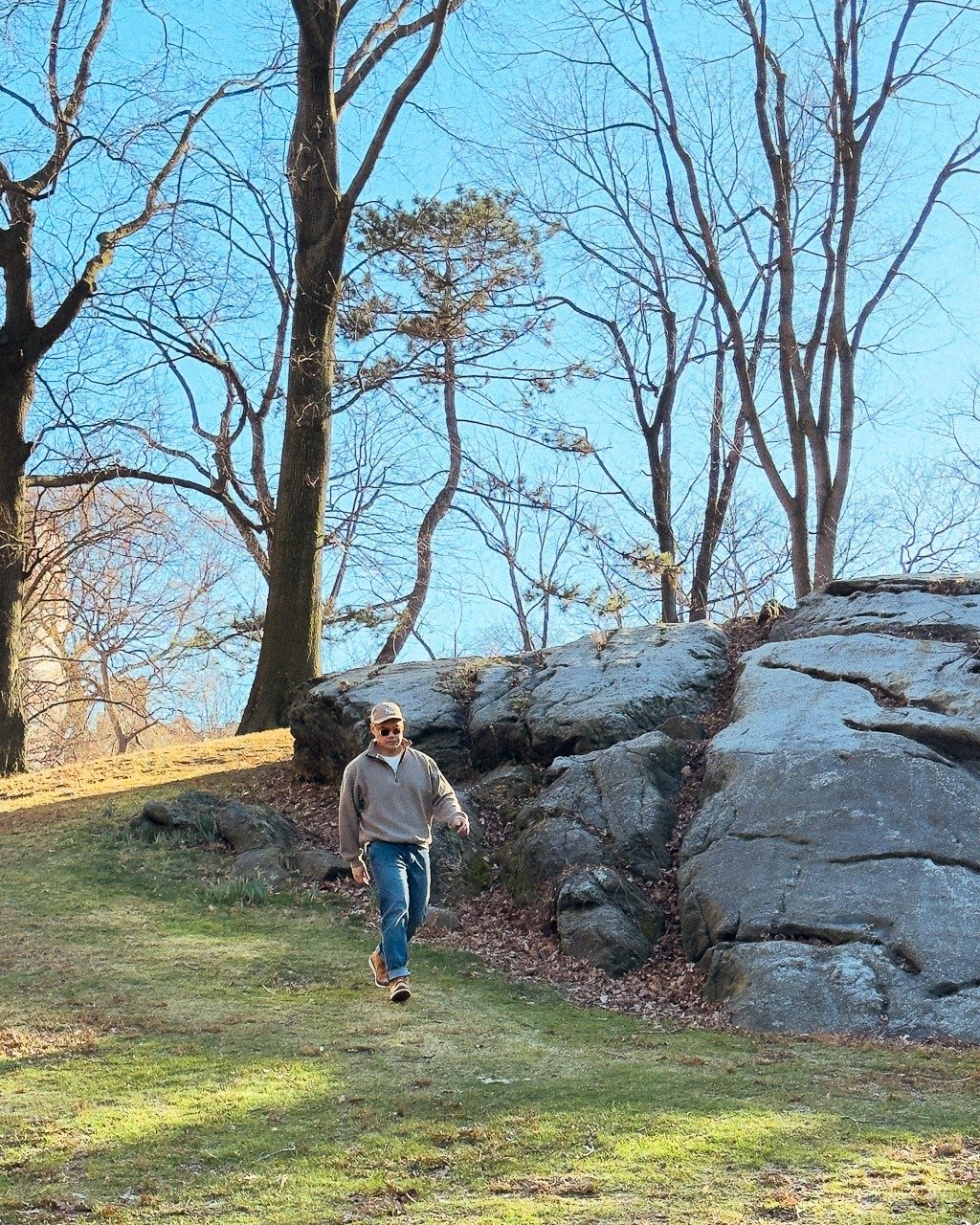 homme en jeans et sweat-shirt à fermeture éclair et casquette marchant sur une colline d'herbe verte dans un parc avec des rochers et de grands arbres en arrière-plan