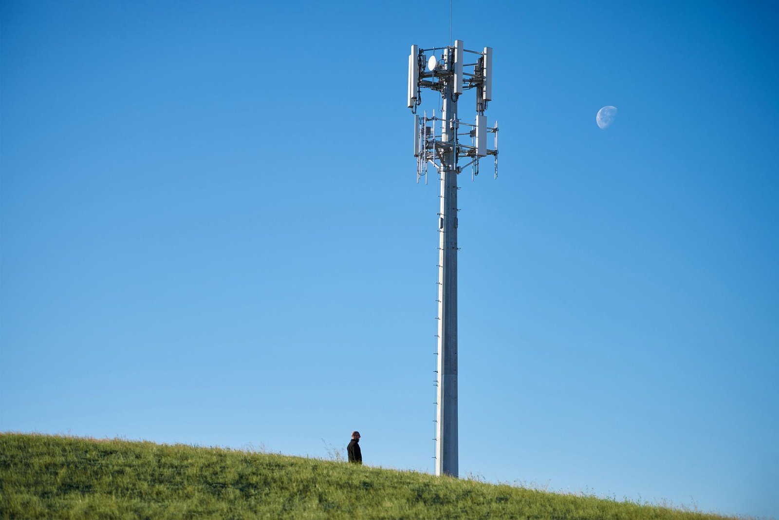 Tour cellulaire sur une colline avec une personne debout en contrebas et une lune diurne dans le ciel.