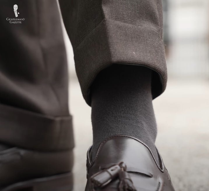 Gros plan des pantalons bruns de Jonas Froelich Hiltner avec ourlets à menace, chaussettes grises et mocassins de gland brun foncé