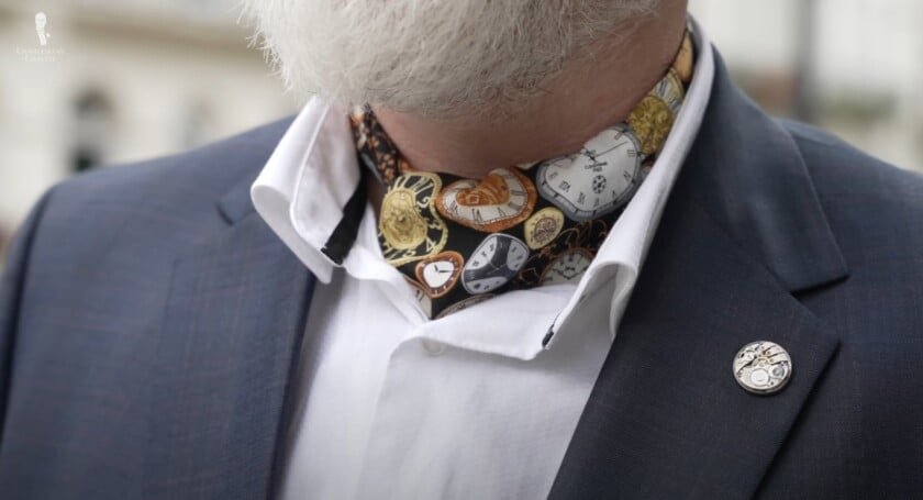 Un gros plan des cravates d'un gentleman avec une écharpe en soie avec une horloge complexe et des imprimés pour le visage de montre, associés à une épingle de revers de montre mécanique sur un blazer bleu marine