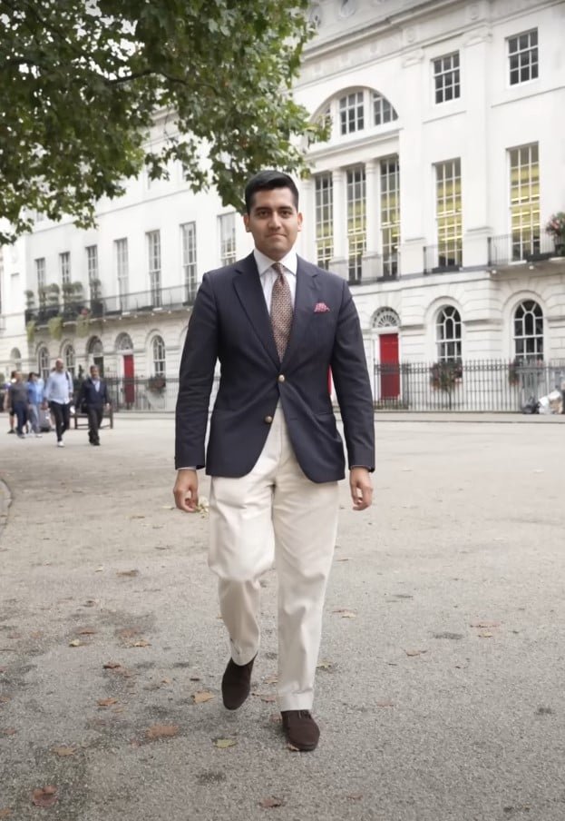 Antonio du Pérou portant un blazer bleu marine avec des boutons en or, une chemise sur mesure blanche de Vestitificio à Milan, une cravate Burberry et un pantalon sur mesure crème de Marco Serrato.