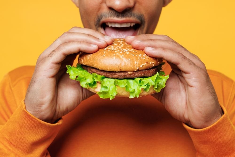 Le jeune homme cultivé grossit porte des vêtements décontractés oranges tenant et mangeant un hamburger.