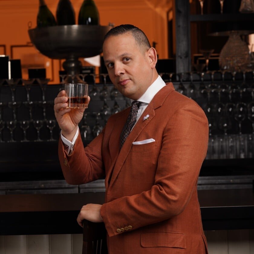 Lookbook d'automne 2022 Raphael Rust Orange Suit Cropped Square Raphael se tient dans un bar portant un costume d'orange rouillé tenant un verre