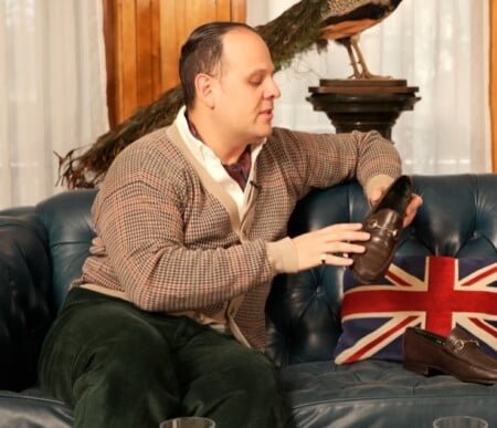 Raphael dans un cardigan de style vintage et un pantalon en velours côtelé vert tient un mocassin de gucci marron tout en étant assis sur un canapé en cuir bleu