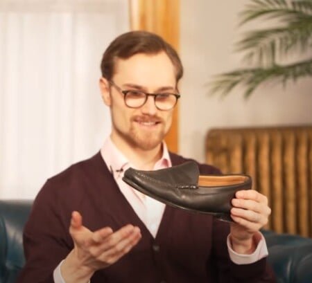 Preston dans des verres et un cardigan bordeaux sourit tout en tenant un mocassin noir de majordome noir, assis sur un canapé en cuir bleu
