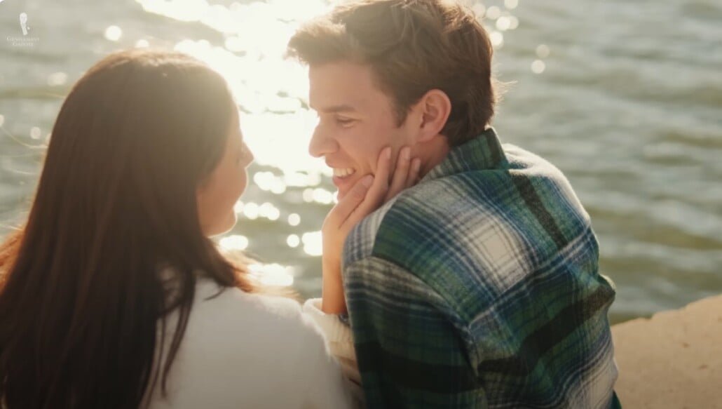 Gentleman vice philandering Jeune couple assis près de l'eau au coucher du soleil, souriant alors que la femme touche doucement le visage de l'homme pendant qu'ils se regardent affectueusement