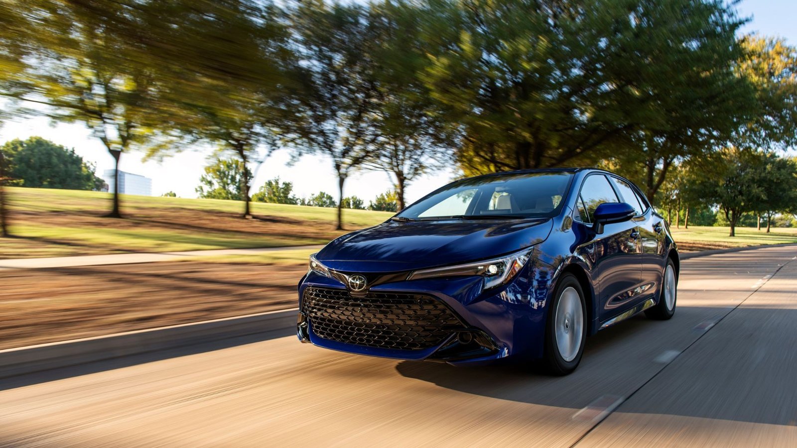 Dynamic Front 3/4 THOT ONE BLUE 2025 TOYOTA COROLLA HATCHBACK SE conduisant sur une route recouverte d'arbres.