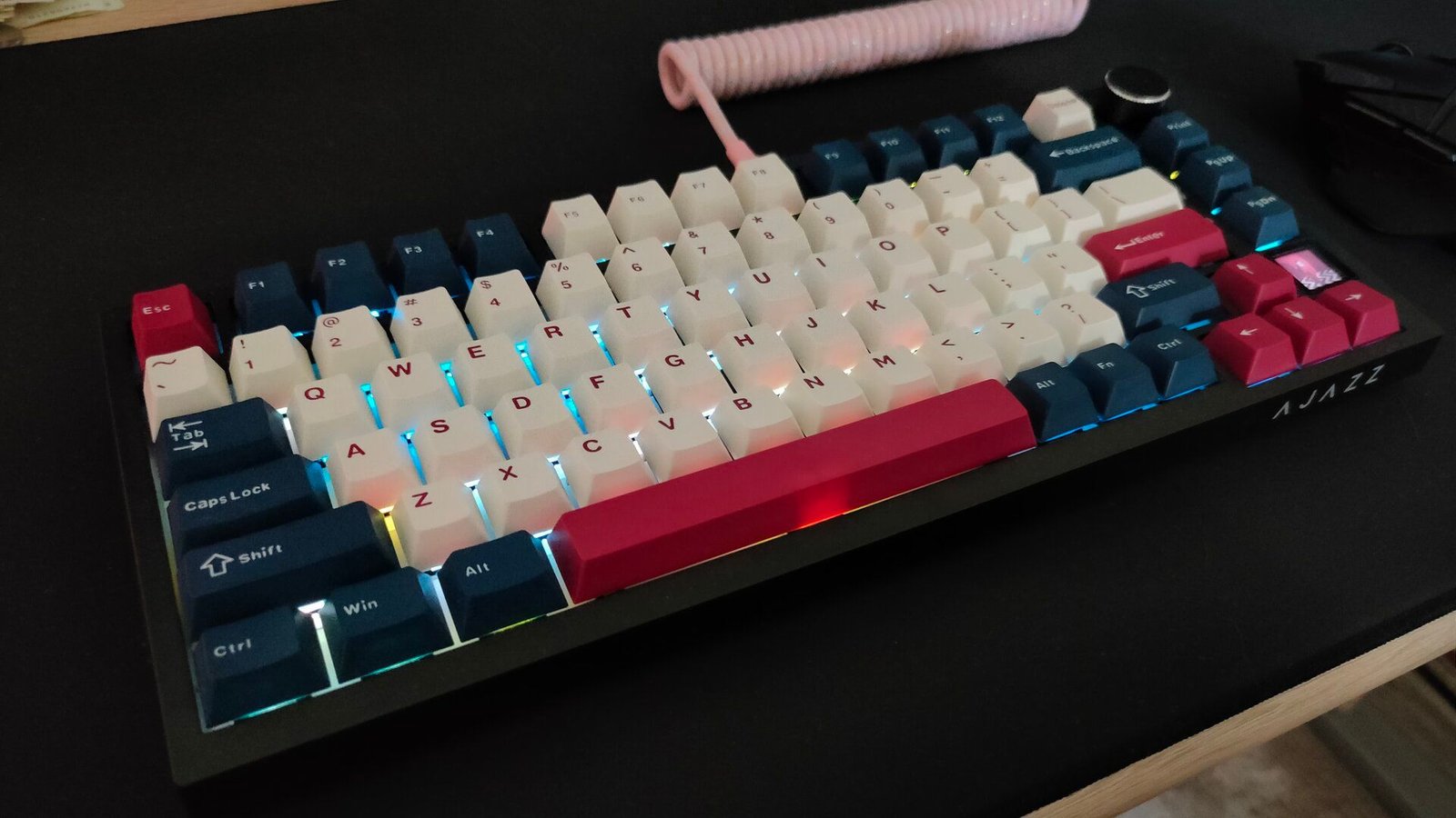 Le clavier mécanique Ajazz AK820 Pro avec un câble enroulé rose et des touchés blancs, bleus et rouges assis sur un bureau avec une souris de soute de SteelSeries.
