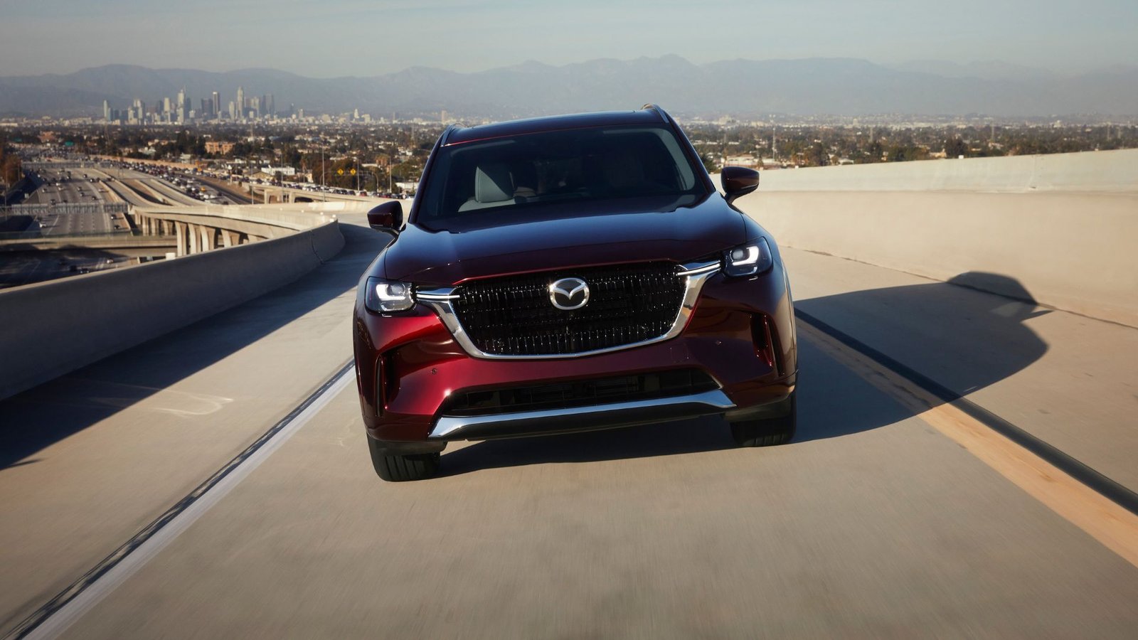 Front de front dynamique d'une Mazda CX-90 rouge 2025 conduisant sur une autoroute avec un paysage urbain en arrière-plan.