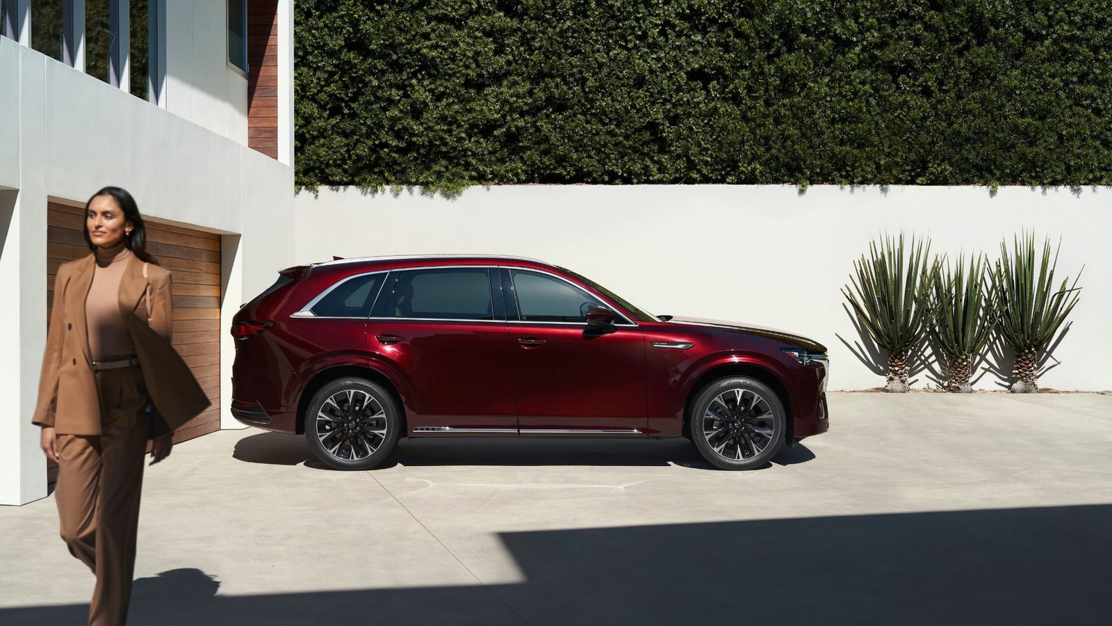 Profil latéral statique CHAMBRE D'UNE MAZDA CX-90 ROUGE 2025 GADÉE SUR UNE RÉDUCTION avec une femme marchant au premier plan.