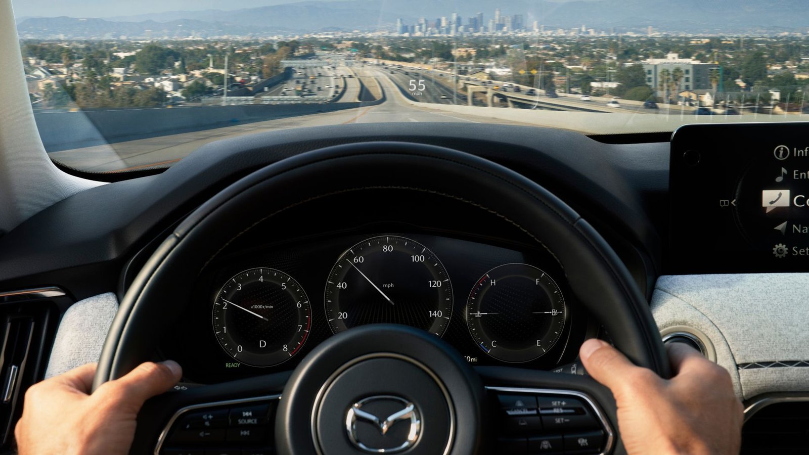 Close-up tir des mains d'un homme sur le volant d'une Mazda CX-90 en 2025, avec un Citscape en arrière-plan.