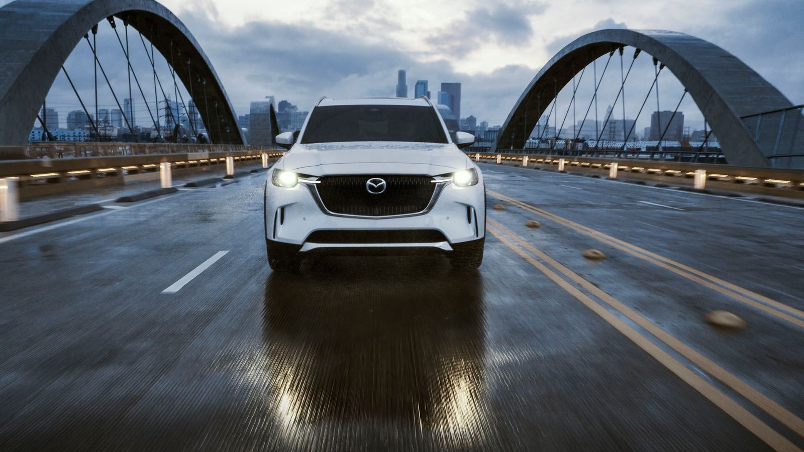 Dynamique Front-end d'une Mazda CX-90 blanche en 2025 conduisant sur un pont avec un paysage urbain en arrière-plan.
