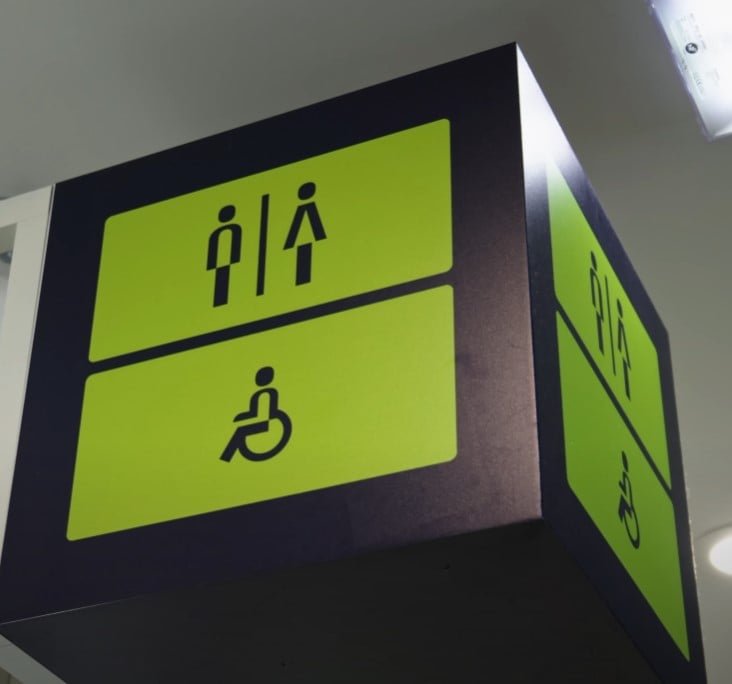 Signe aérien avec fond vert et icônes noires indiquant des toilettes masculines et féminines au-dessus d'un symbole pour les installations accessibles en fauteuil roulant.