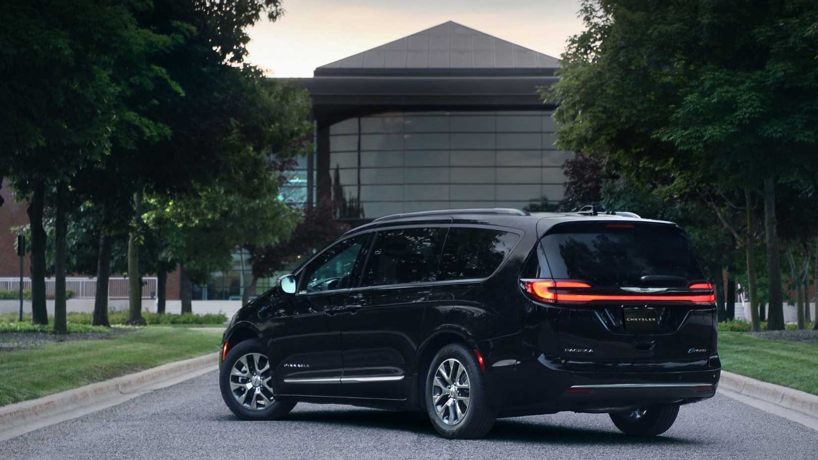 Arrière statique 3/4 THOT ONE Black 2025 Chrysler Pacifica Plug-in Hybrid Pinnacle avec un bâtiment moderne et des arbres en arrière-plan.
