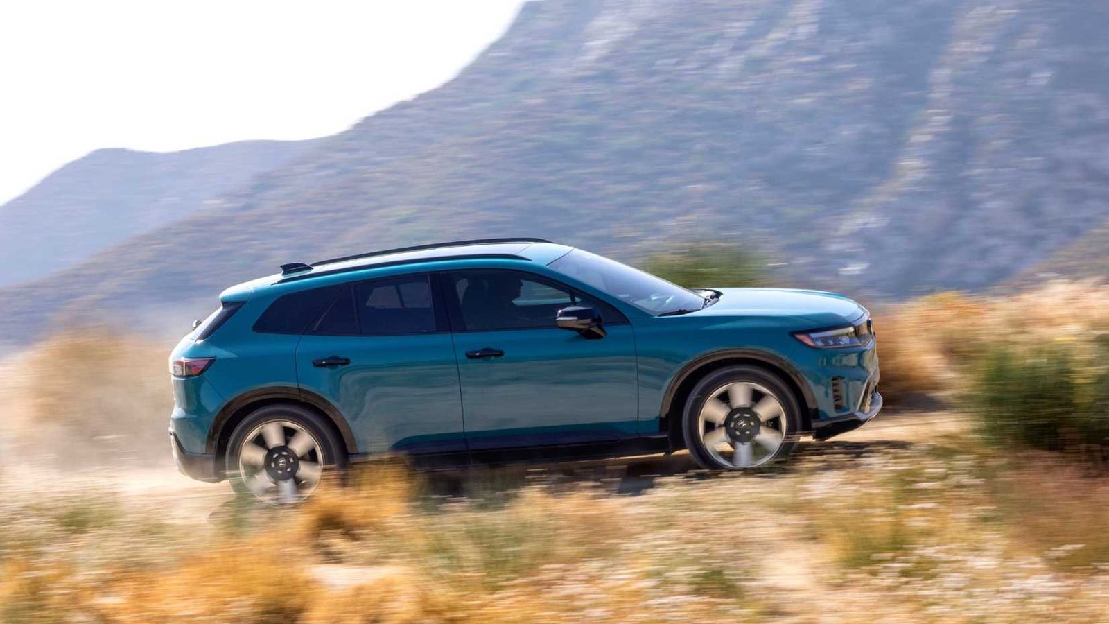 Profil latéral dynamique THOT d'une Honda Prologue Blue 2025 conduisant sur une piste de terre avec des montagnes en arrière-plan.