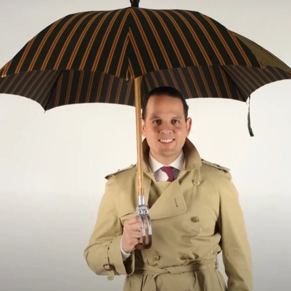 Un homme souriant dans un trench-coat tenant un parapluie rayé ouvert.