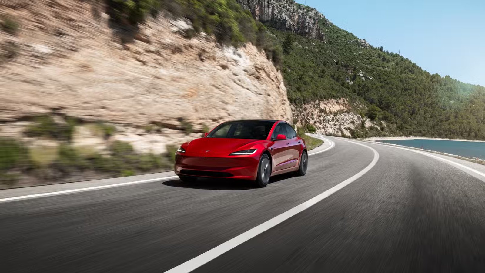 Plan dynamique 3/4 avant d'une Tesla Model 3 rouge roulant sur une route côtière.