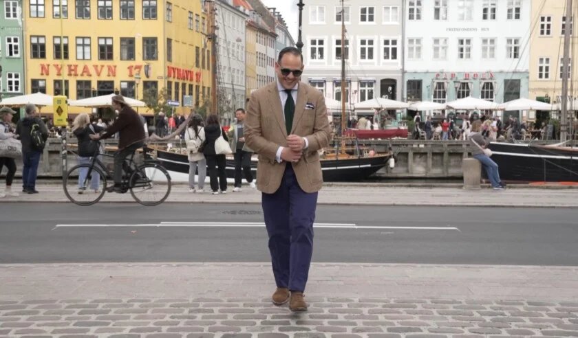 Raphaël dans un manteau de sport en tweed beige, une cravate verte, un pantalon bleu et des chaussures en daim marron se promène le long du port de Nyhavn à Copenhague.