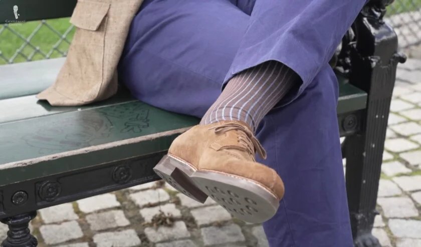 Vue rapprochée des jambes croisées d'un homme, montrant un pantalon bleu, des chaussettes rayées marron et bleu clair et des chaussures derby en daim marron avec d'épaisses semelles en caoutchouc.