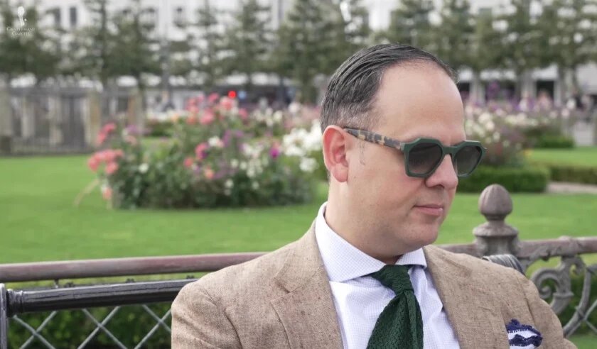 Gros plan de Raphael portant une veste en tweed beige, une cravate en tricot verte et des lunettes de soleil à monture verte distinctives, assis sur un banc de parc.