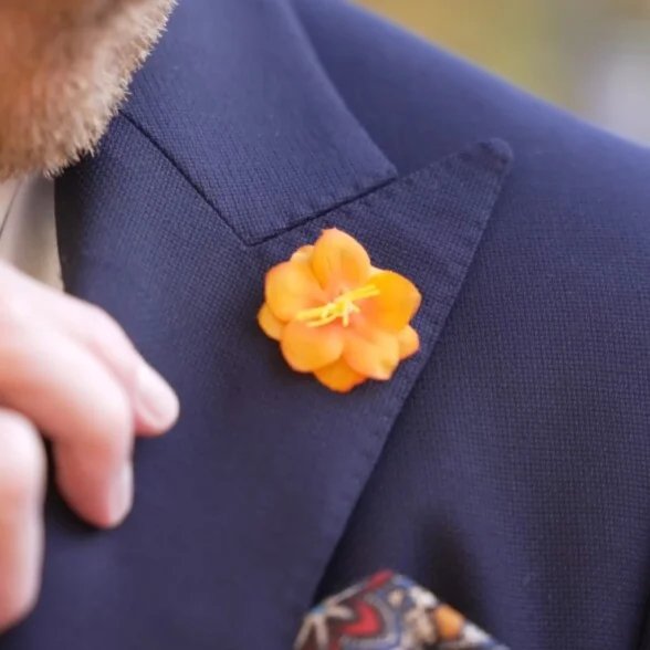 La main de Preston ajustant une boutonnière en soie Fort Belvedere orange vif sur le revers d'une veste de costume bleu marine