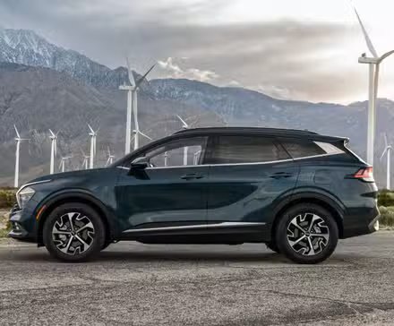 Photo de profil latérale statique d'une Kia Sportage hybride 2025 grise avec des montagnes et des éoliennes en arrière-plan.