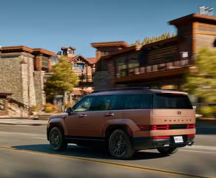 Plan dynamique 3/4 arrière d'une Hyundai Santa Fe hybride 2025 en bronze traversant une ville.