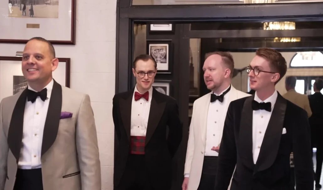 Quatre messieurs vêtus de tenues variées Black Tie, notamment des smokings et des smokings blancs et noirs, arrivent ensemble sur un lieu d'événement officiel.