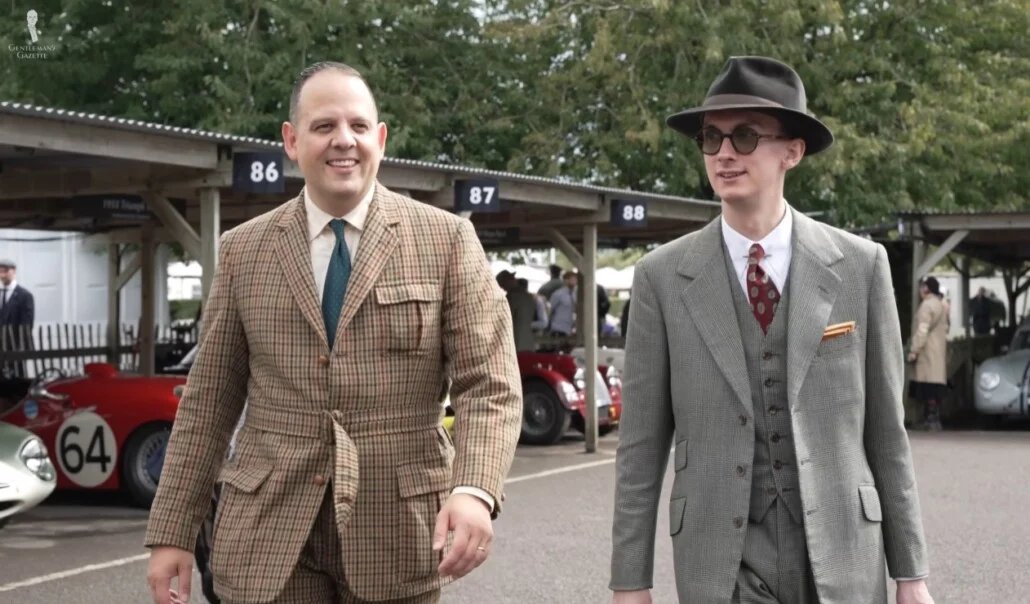 Deux messieurs portant des costumes en tweed d'inspiration vintage, un fedora et des lunettes de soleil, se promenant dans le paddock d'un salon de voitures classiques avec des véhicules de course vintage.