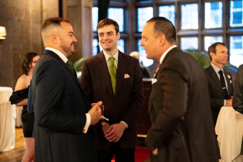 Trois messieurs vêtus de vestes et de cravates classiques pour hommes, souriant et conversant lors d'une réunion sociale.