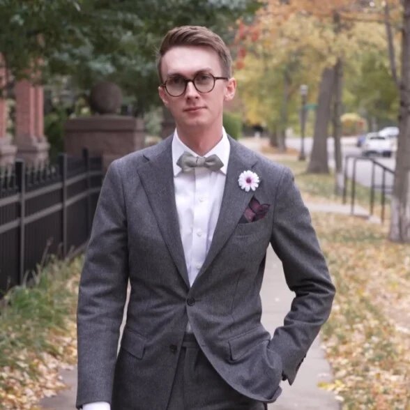 Un gentleman marchant avec confiance dans une rue résidentielle, vêtu d'un costume de flanelle grise, d'un nœud papillon et d'une boutonnière.