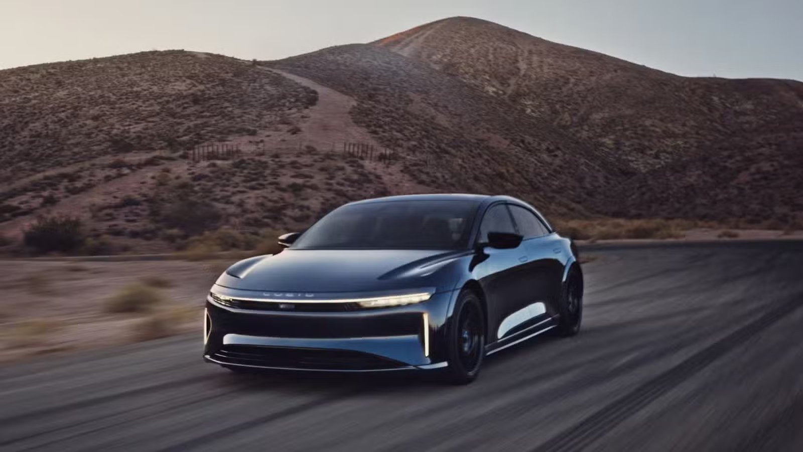 Vue complète d'un Lucid Air Sapphire bleu roulant sur une route de campagne avec une montagne en arrière-plan.