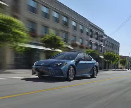 Vue 3/4 avant d'une Toyota Camry 2025 bleue traversant une ville.