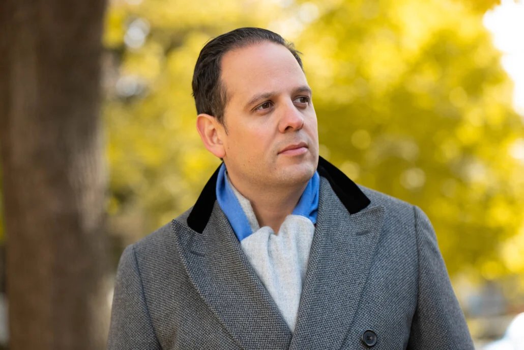Raphaël porte un pardessus gris à double boutonnage avec un col en velours noir, associé à une écharpe unie bicolore bleu clair et gris sur un fond automnal.