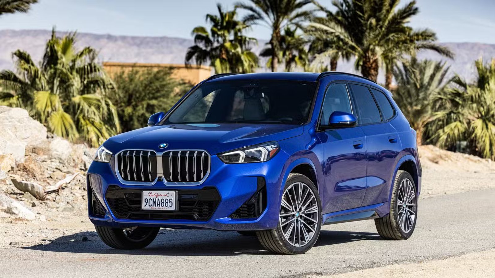 Plan statique 3/4 avant d'une BMW X1 xDrive28i 2023 bleue avec des palmiers en arrière-plan.