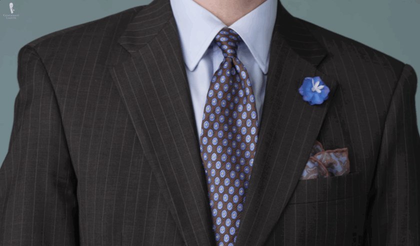 Gros plan d'un revers de costume marron anthracite avec une boutonnière en delphinium bleu, une cravate vintage marron et une pochette de costume motif cachemire bleu et marron.