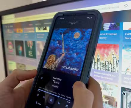 Les mains d'une personne tenant un téléphone Anrdoid avec l'application Finamp devant un écran d'ordinateur.