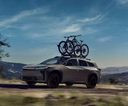 Plan dynamique 3/4 avant d'une Toyota bZ Woodland 2026 avec des vélos de montagne sur son toit roulant sur un chemin de terre avec des montagnes en arrière-plan.