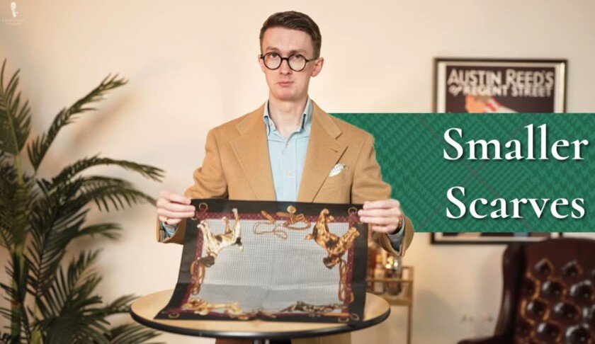 Options de style pour un petit foulard en soie 1 Homme vêtu d'un manteau de sport camel tenant un petit foulard carré en soie au-dessus d'une table, avec le texte à l'écran indiquant « Petits foulards"