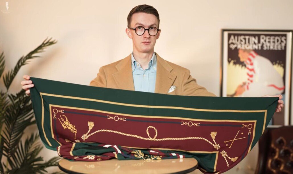 Guide de style pour les grands foulards en soie Homme dans un manteau de sport camel tenant un grand foulard carré en soie avec une bordure verte et un centre à motifs bordeaux