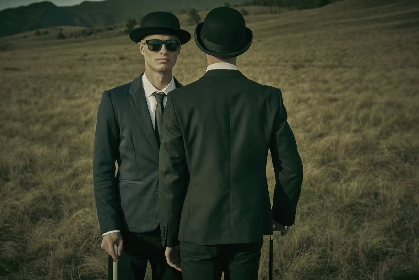 Deux hommes sérieux et calmes en costumes noirs, chapeaux melon et lunettes de soleil noires se font face dans un champ sauvage ouvert.