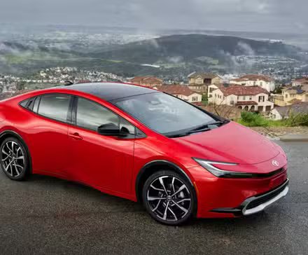 Plan statique 3/4 avant d'une Toyota Prius PHEV XSE 2025 rouge avec des nuages ​​et une ville en arrière-plan.