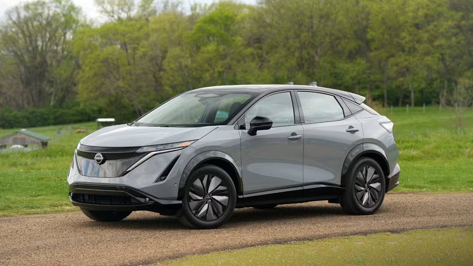 Vue 3/4 avant d'une Nissan Ariya 2025 grise garée sur une piste de gravier avec de l'herbe et des arbres en arrière-plan.