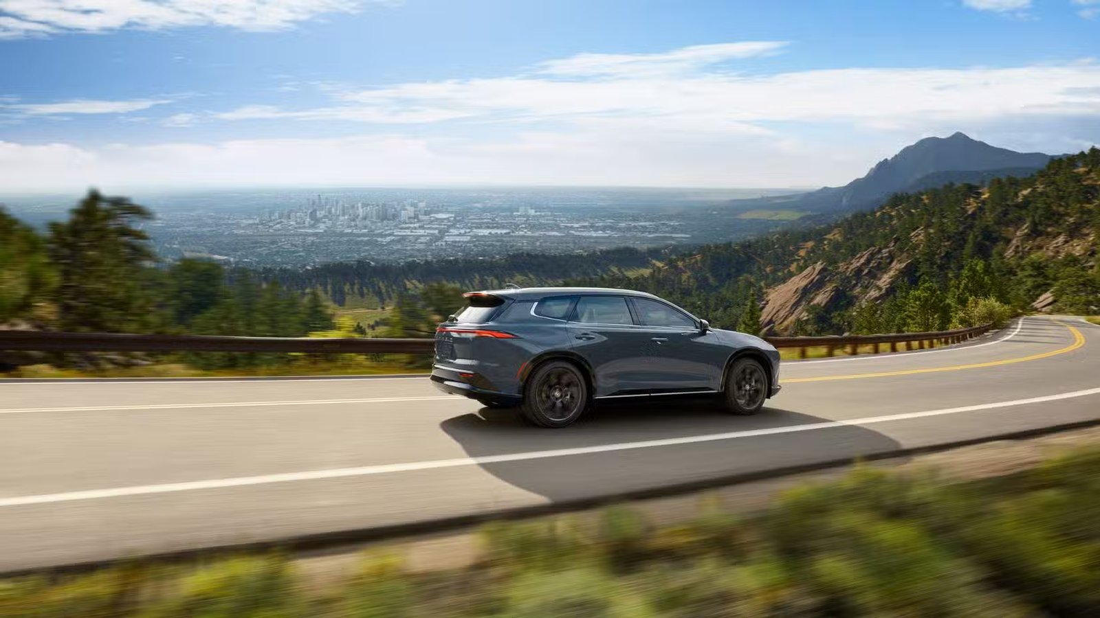 Photo de profil latérale dynamique d'une Toyota Crown Signia 2025 grise roulant sur une route de campagne à travers les montagnes avec une ville au loin.