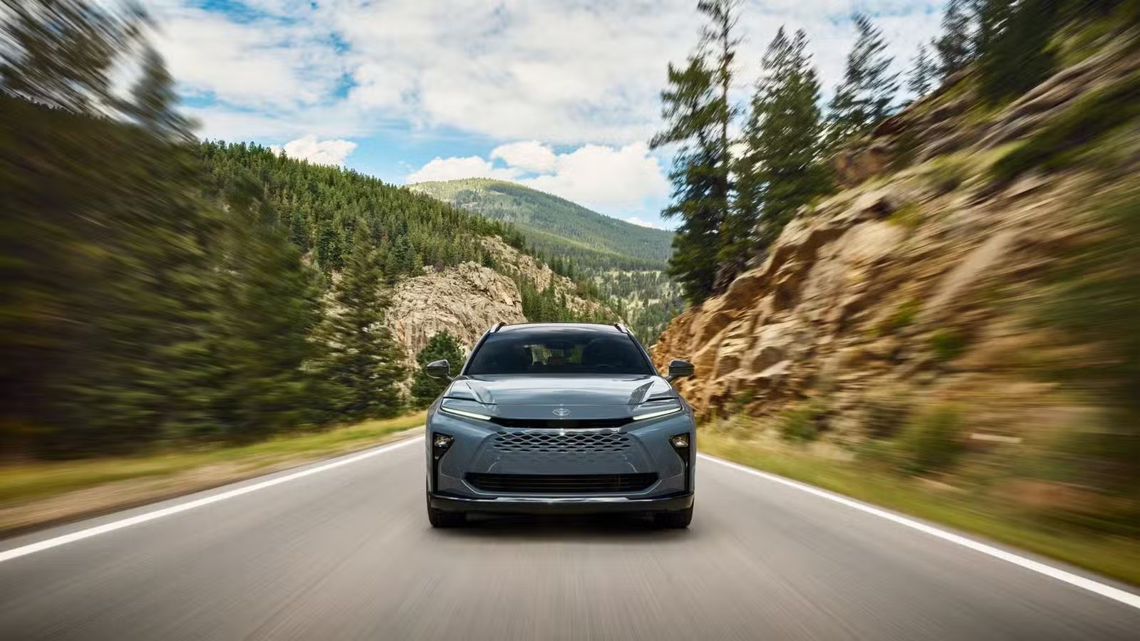 Plan avant dynamique d'une Toyota Crown Signia 2025 grise roulant sur une route de campagne bordée d'arbres et d'une montagne en arrière-plan.