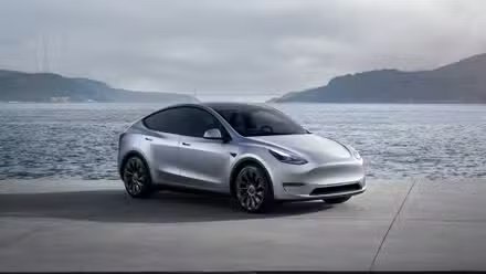 Une Tesla Model Y argentée garée sur le tarmac avec des montagnes et une baie d'eau derrière elle.